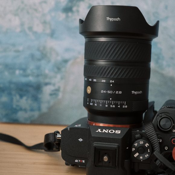 A Sony camera with a Thypoch 24-50mm lens sits on a wooden surface against a blue background.