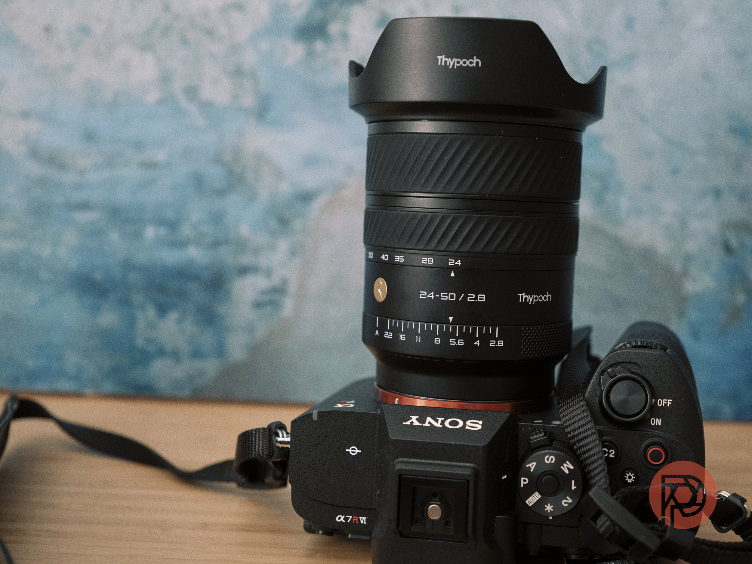 A Sony camera with a Thypoch 24-50mm lens sits on a wooden surface against a blue background.