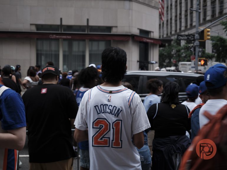 A crowd gathers on a city street; a person in a Dotson 21 jersey stands in the foreground.