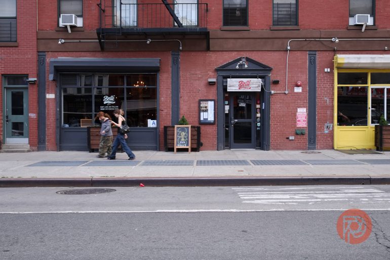 Two people walk past a red brick restaurant on a city street with storefronts and a sidewalk.