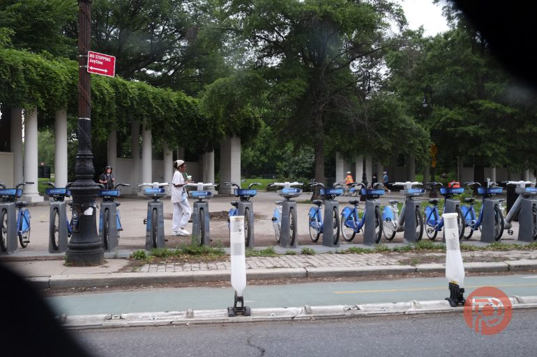 A row of blue bikes at a bike share station with people walking by and trees in the background.