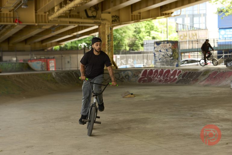 A man rides a BMX bike in a graffiti-covered skatepark under a bridge.