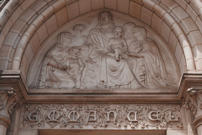Stone relief of five adults and two children beneath an arched entryway with decorative text below.