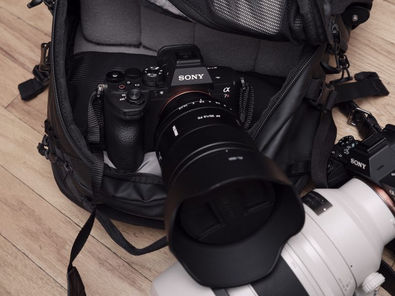 A Sony camera with a lens sits in an open camera bag next to another camera with a large white lens.