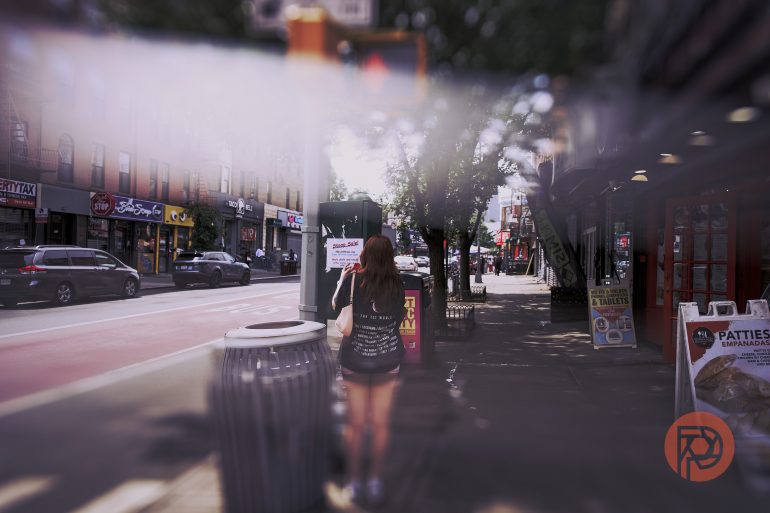 A woman stands on a city sidewalk near shops and a trash bin, with cars parked along the street.
