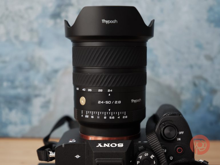 Close-up of a Thypoch 24-50mm camera lens mounted on a Sony camera against a blurred blue background.