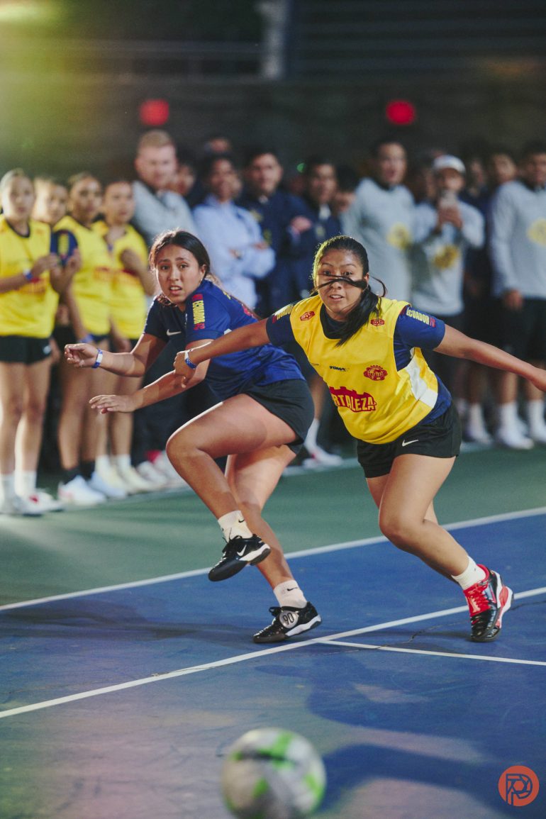 Two female players chase a soccer ball on an indoor court, with teams and spectators watching in the background.