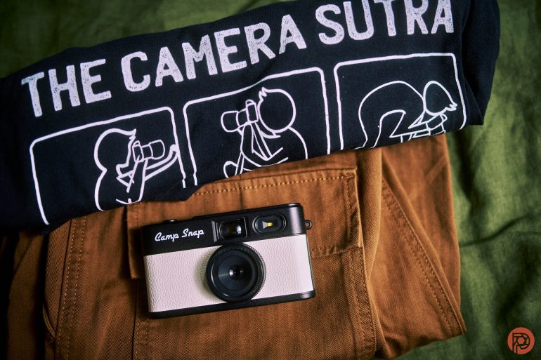 A t-shirt with The Camera Sutra illustrations, folded above brown pants, with a vintage camera on top.
