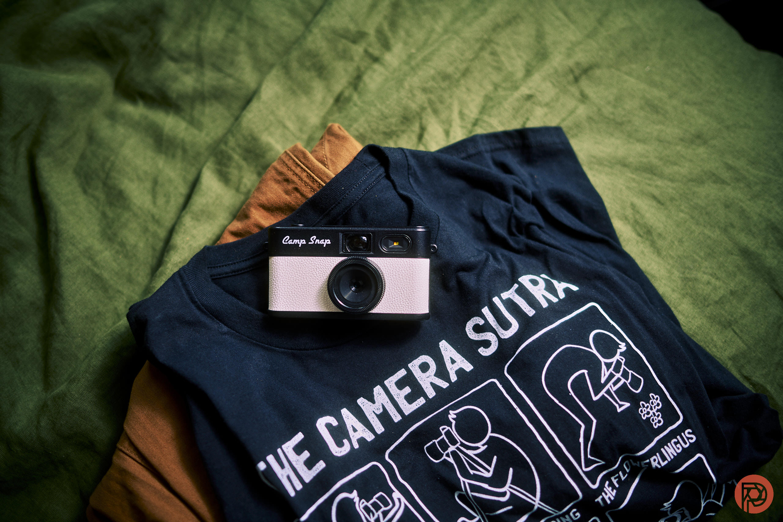 Camp Snap 2 Review: They Did the Right Thing A vintage camera on a black graphic T-shirt, both laid out on a green fabric background.