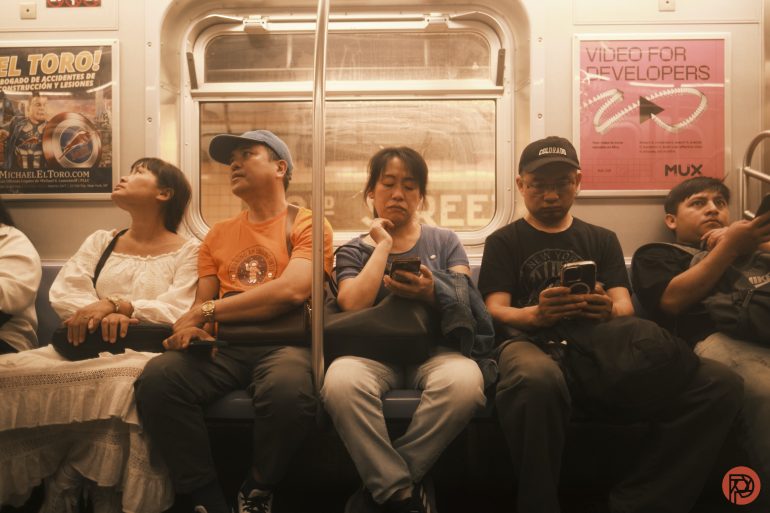 Five people sitting on a subway bench, four looking at phones, one gazing up; advertisements in the background.