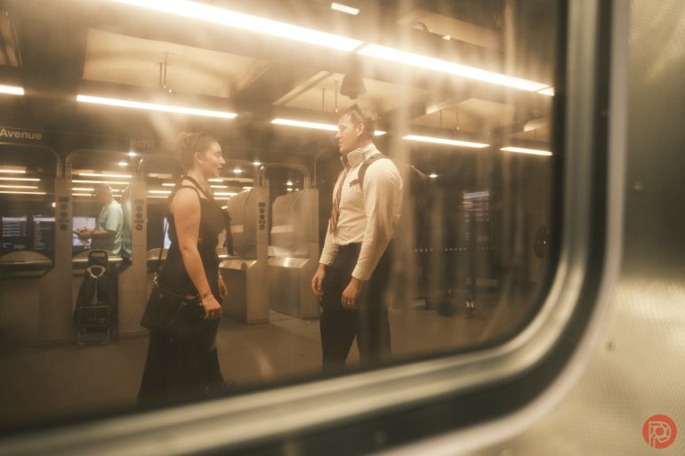 Two people stand and talk on a subway platform, seen through a window with reflections and warm lighting.