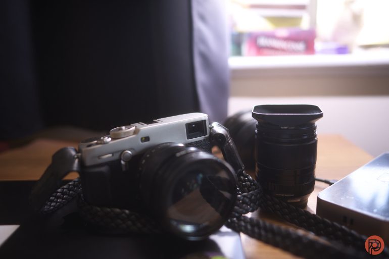 A camera with a braided strap and a separate lens sit on a wooden desk near a window.