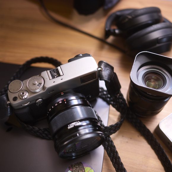 A camera, an extra lens, and headphones on a wooden desk with a black laptop and a mouse.