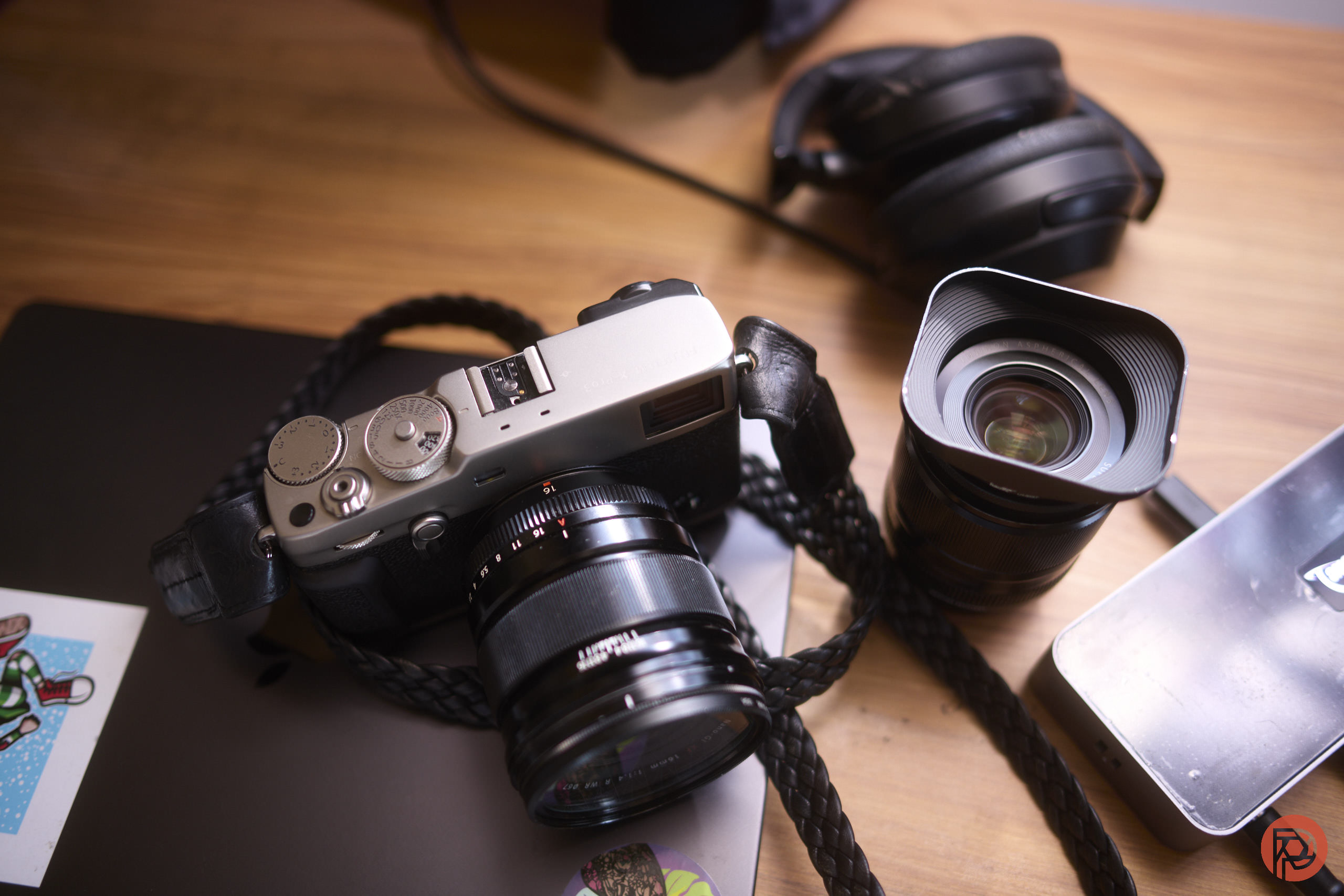 How a Bunch of Fanboys Ruined Fujifilm A camera, an extra lens, and headphones on a wooden desk with a black laptop and a mouse.