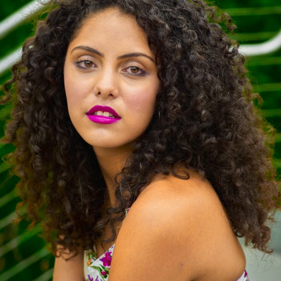 Woman with curly hair and bright pink lipstick sits outdoors, wearing a floral top, looking at the camera.