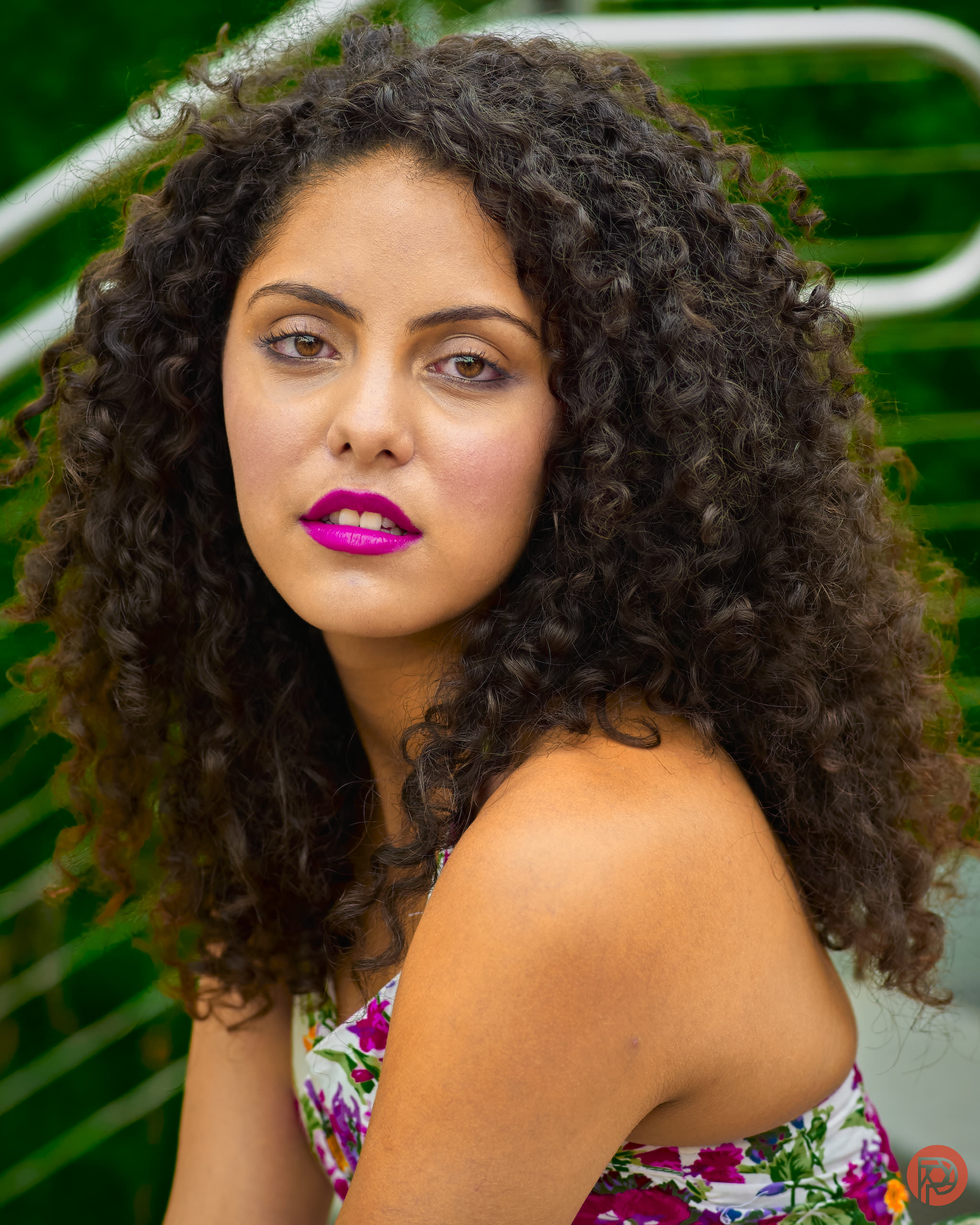 Woman with curly hair and bright pink lipstick sits outdoors, wearing a floral top, looking at the camera.