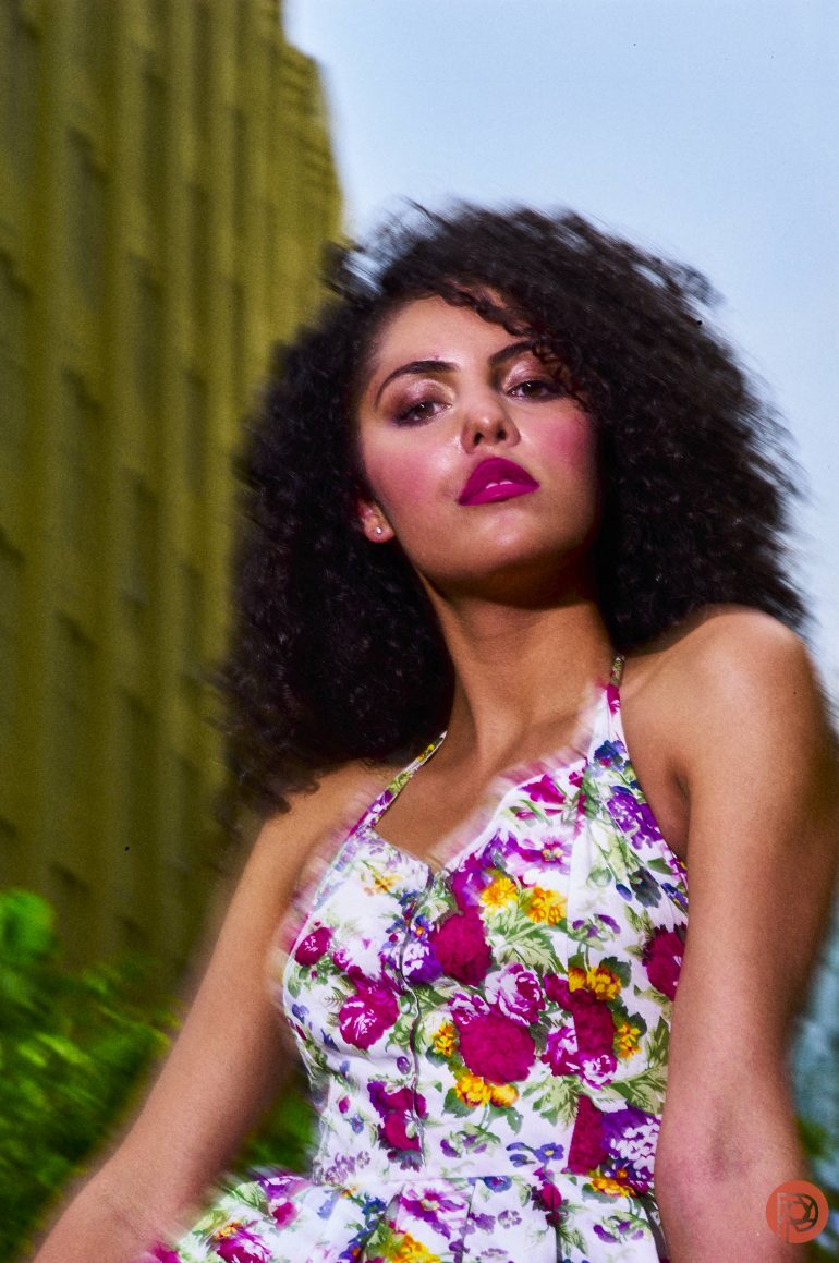 Woman with curly hair in a floral dress poses confidently outdoors beside a tall yellow building.
