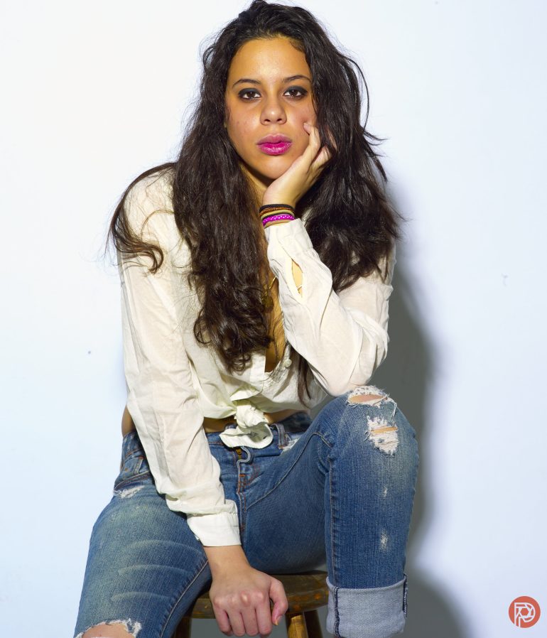 Woman with long dark hair in ripped jeans and a white shirt sits on a stool against a plain background.