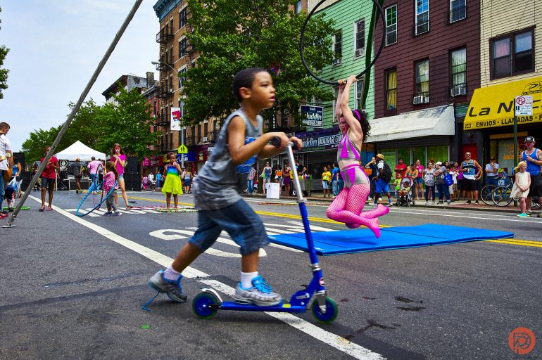 A boy rides a scooter on a city street as a performer swings on a hoop, watched by a crowd.