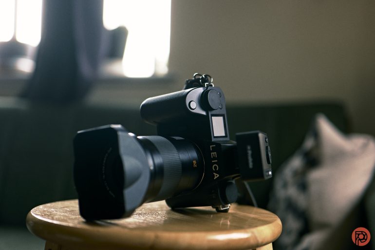 A Leica camera rests on a wooden stool, with a sofa and pillow blurred in the background.