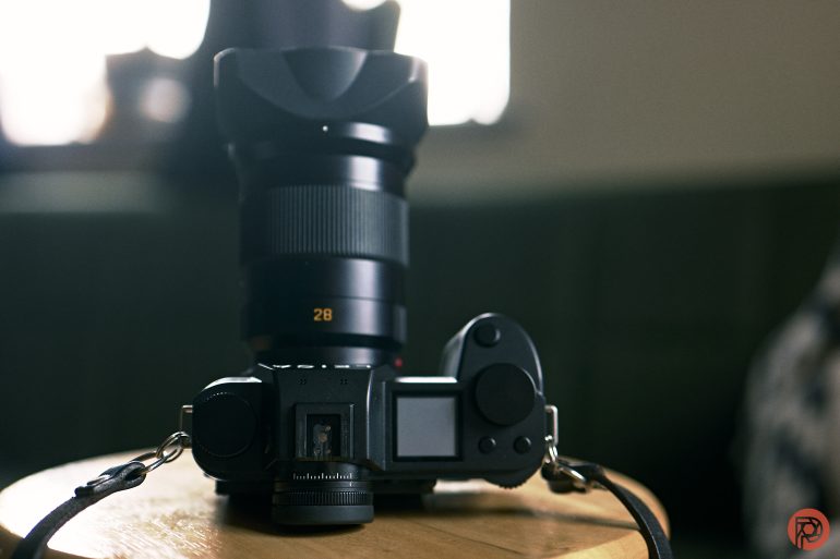 A digital camera with a large lens rests on a wooden stool in a softly lit room.