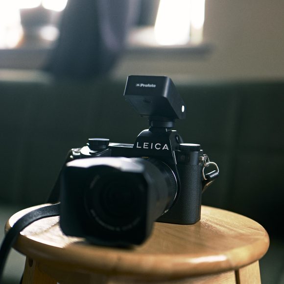A Leica camera with a Profoto flash attachment sits on a wooden stool in a dimly lit room.