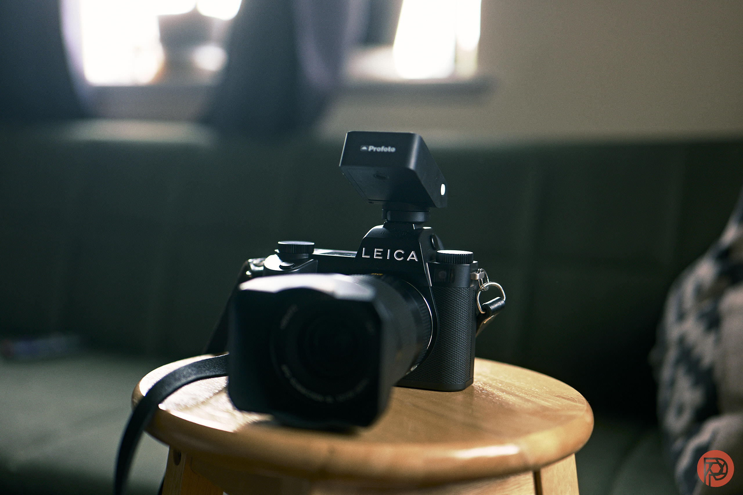 6 Cameras From 2026 Every Photographer Should Have On Their Radar A Leica camera with a Profoto flash attachment sits on a wooden stool in a dimly lit room.