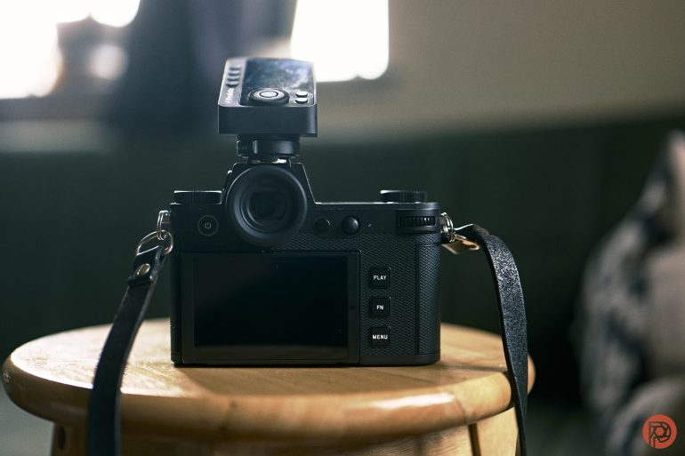 A digital camera with a flash attachment sits on a wooden stool indoors.