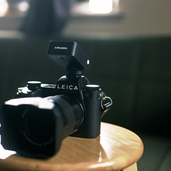 A Leica camera with a Profoto flash sits on a wooden stool in a softly lit room.