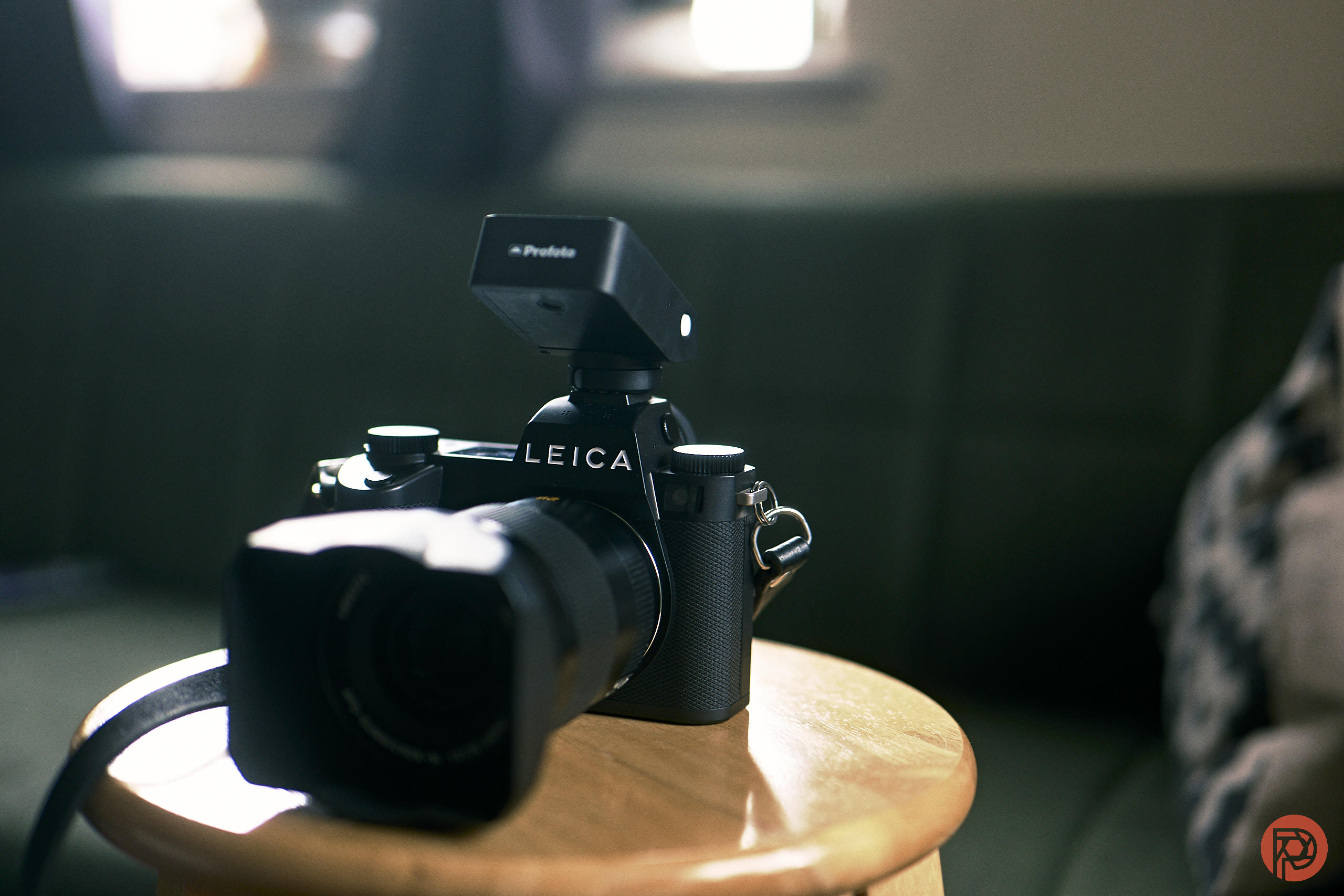 A Leica camera with a Profoto flash sits on a wooden stool in a softly lit room.