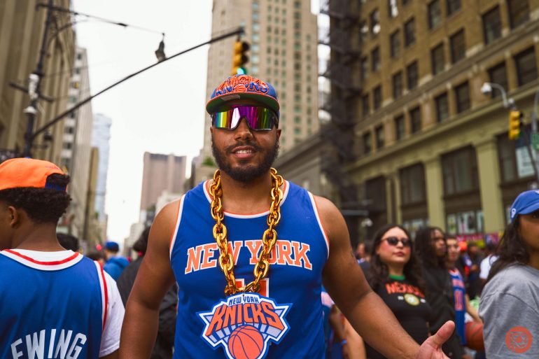 Man in New York Knicks gear and gold chain smiling at a street event, surrounded by other fans.