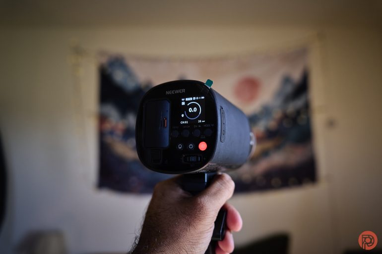 A hand holding a Neewer flash unit, pointed toward a blurred, artistic tapestry background.
