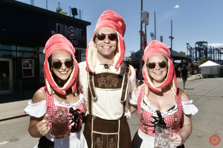 Three people in festive outfits and squid hats smile outdoors, holding beer steins at a street festival.