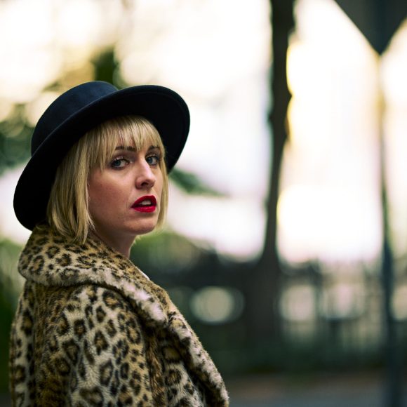 Woman in a leopard print coat and black hat standing outdoors, looking over her shoulder.