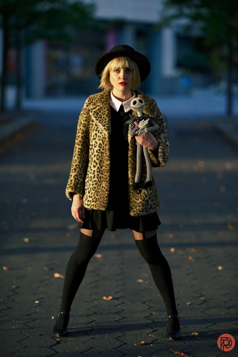 Person in a leopard print coat and black hat holding a Jack Skellington doll stands on a paved path.