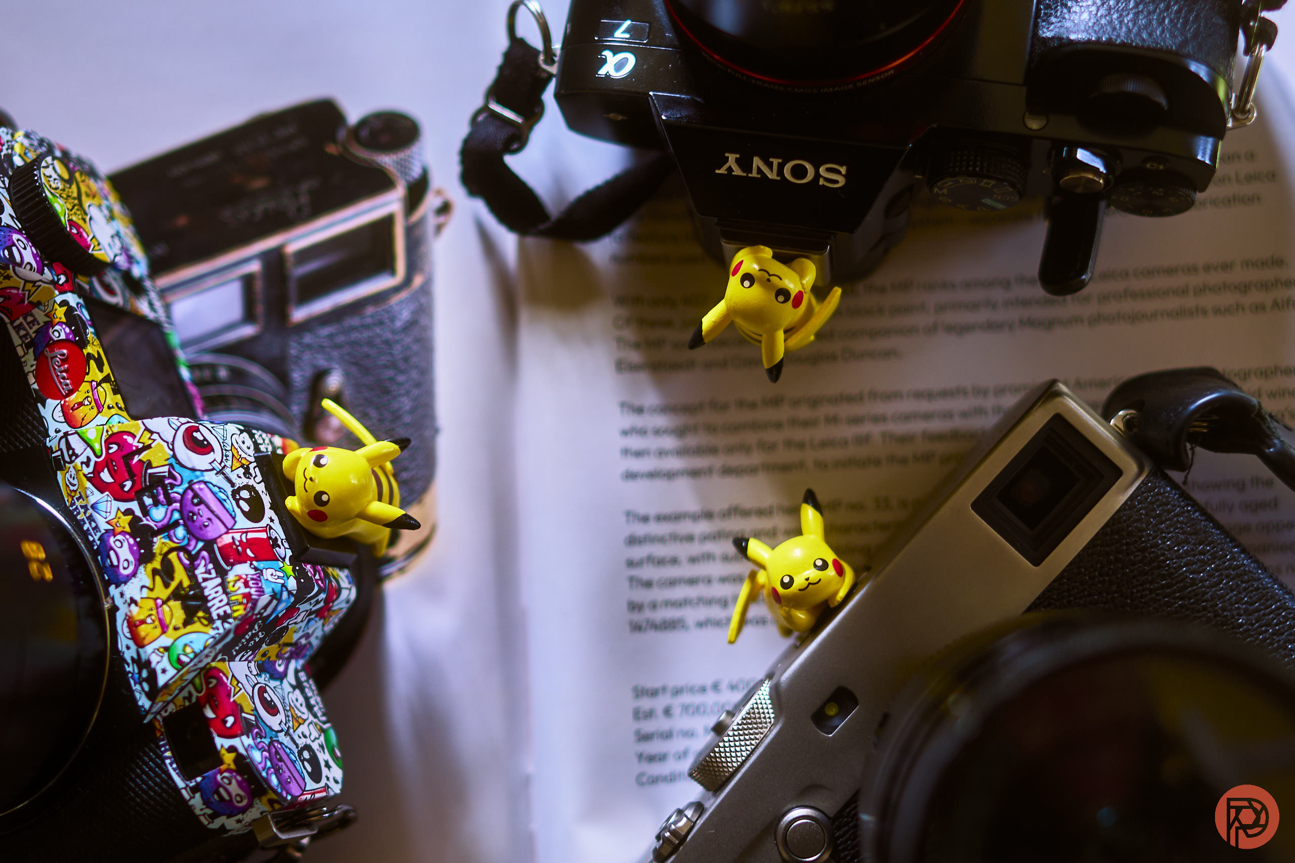 Pikachu Makes For the Very Best Hot Shoe Cover, and I Had to Catch Them All Three Pikachu figurines on vintage cameras, with camera straps and papers in the background.