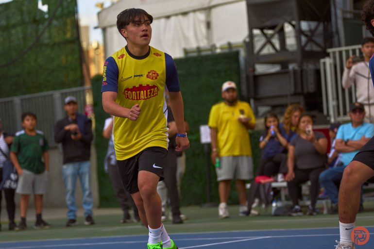 A young athlete in a yellow jersey runs on an outdoor court, with spectators watching in the background.