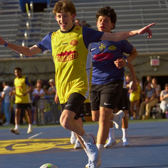 A soccer player in yellow celebrates a goal as others chase him on a blue court, with spectators in the stands.