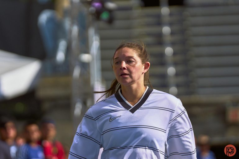 A woman in a white sports jersey stands on a field with stadium seating in the background.