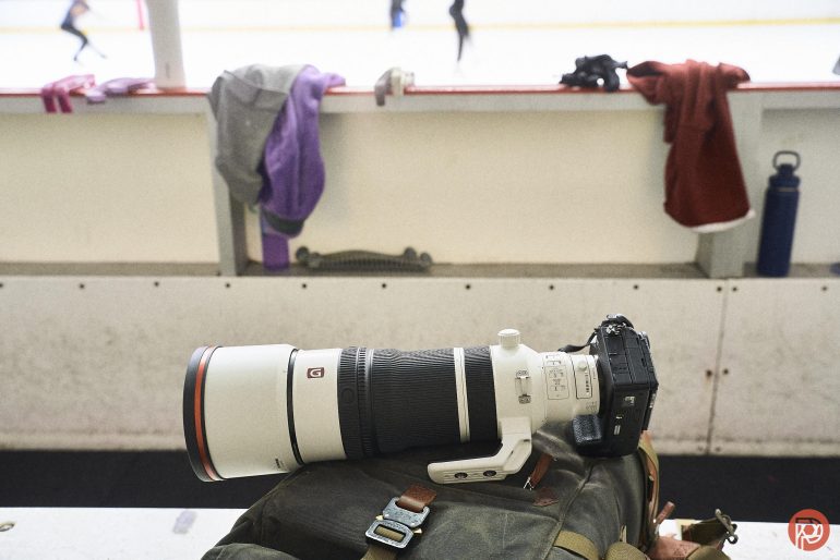 A camera with a large lens rests on a bag at an ice rink, with jerseys hanging on the boards.