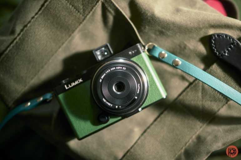 A green Lumix camera with a teal strap rests on an olive green fabric surface.