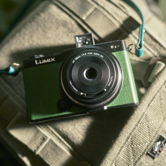 A green Lumix camera with a teal strap resting on a beige fabric bag in sunlight.
