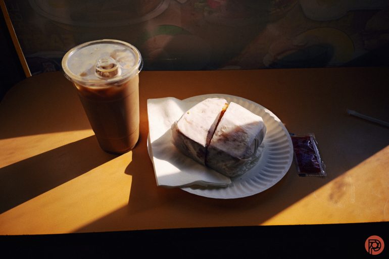 Iced coffee and a wrapped sandwich on a paper plate with napkins, on a sunlit table.