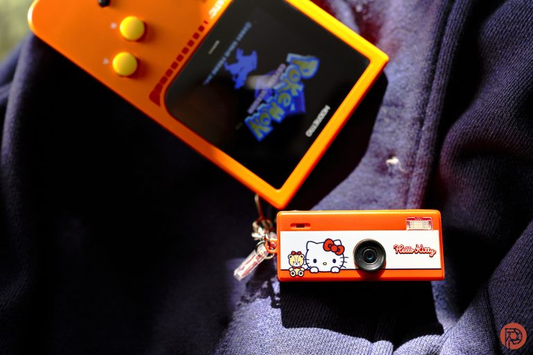 An orange Game Boy with Pokémon on screen and a small orange Hello Kitty camera on a dark fabric background.