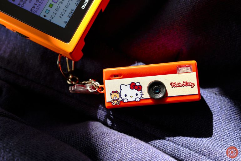 An orange Hello Kitty toy camera on a keychain, resting on dark fabric in bright sunlight.