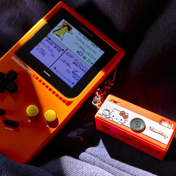 An orange handheld gaming console and a small red Hello Kitty camera on dark fabric in sunlight.