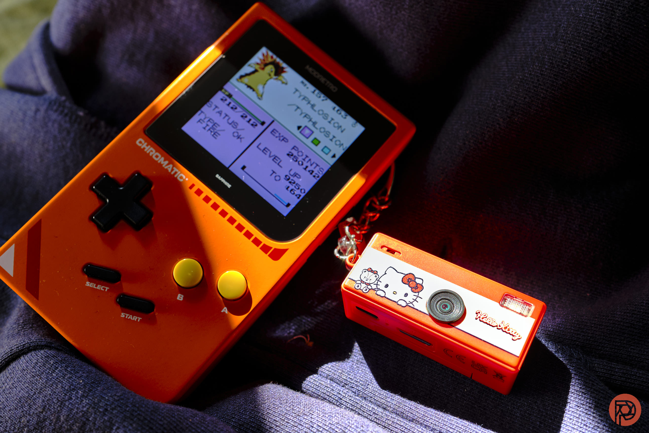 The Yashica Funtastic is a Better Kodak Charmera (Hello Kitty Edition) An orange handheld gaming console and a small red Hello Kitty camera on dark fabric in sunlight.