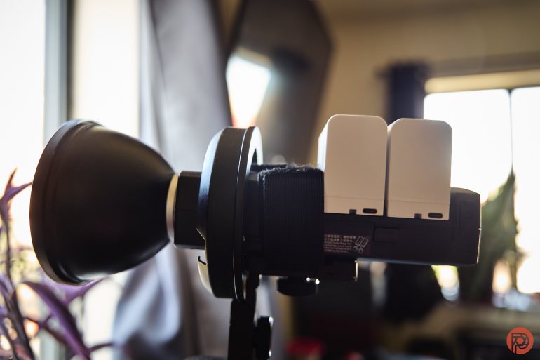 A camera flash unit with two batteries attached, set up indoors near a window with sunlight streaming in.