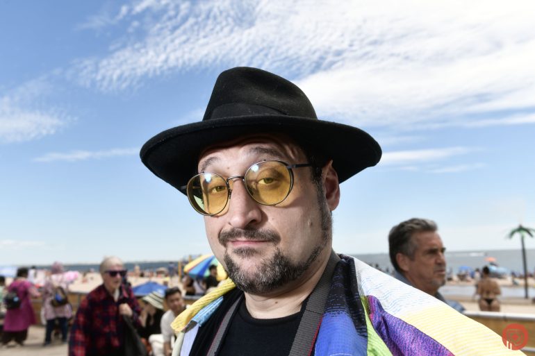 Man in yellow-tinted glasses and black hat at a crowded beach, cloudy sky in background.