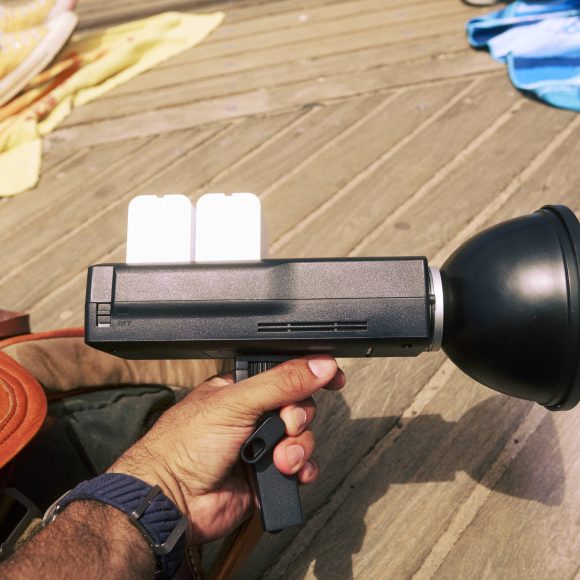 A person holding a handheld black device with a large round front, next to a brown bag on wooden boards.